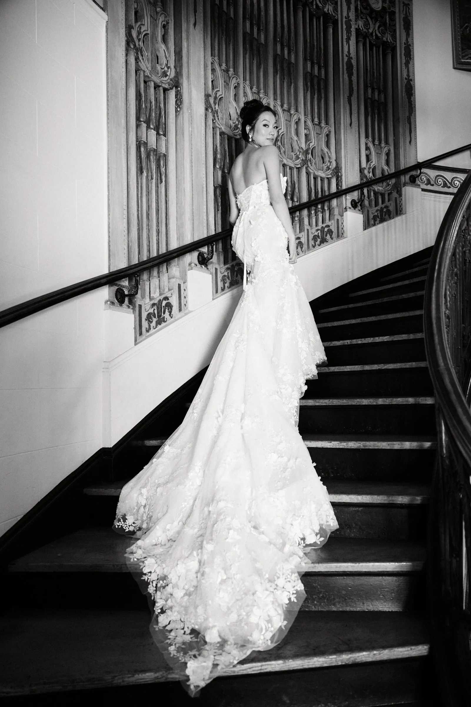 The bride looking back from the mansion's grand staircase, her train trailing down the steps, in black and white