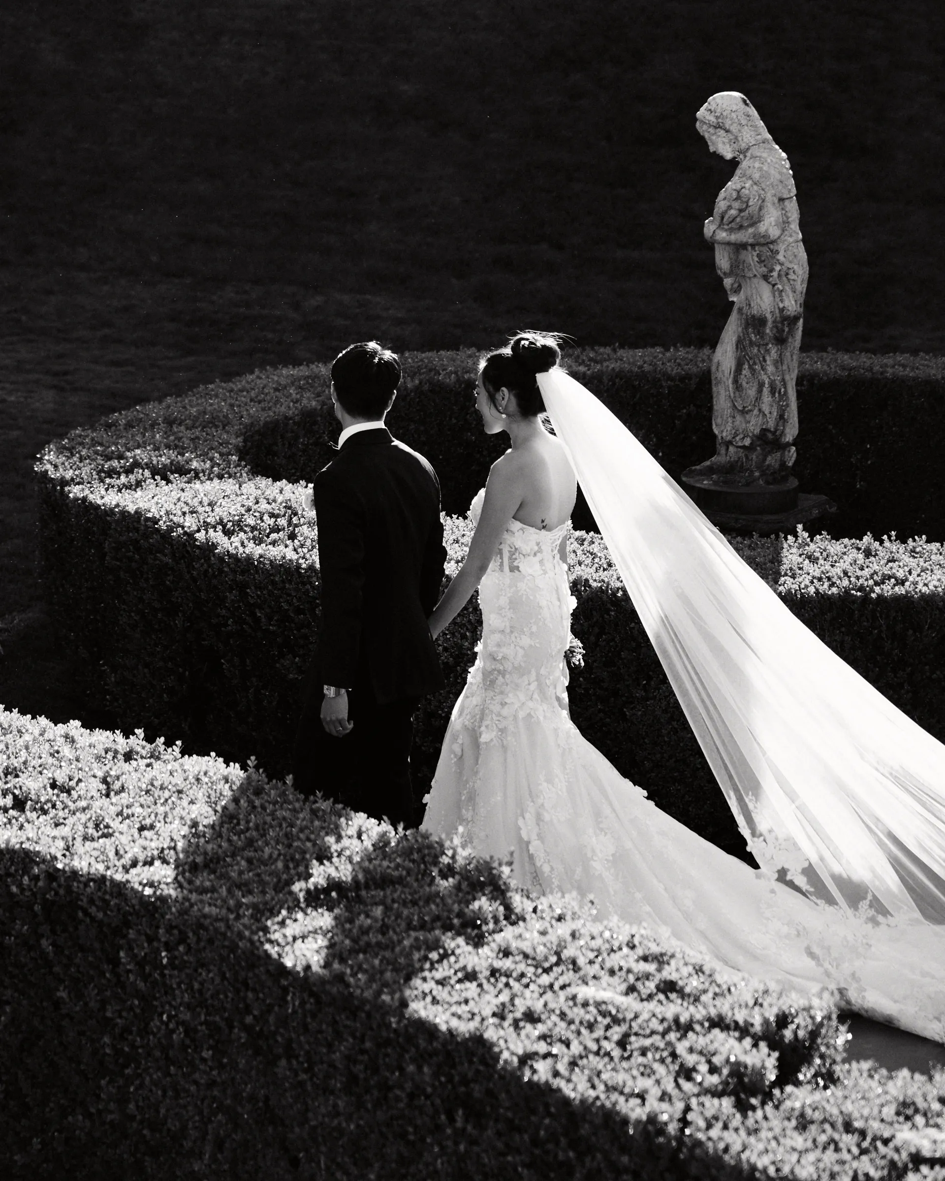 The couple hand in hand in a formal hedge garden, the bride's veil catching the light, in black and white