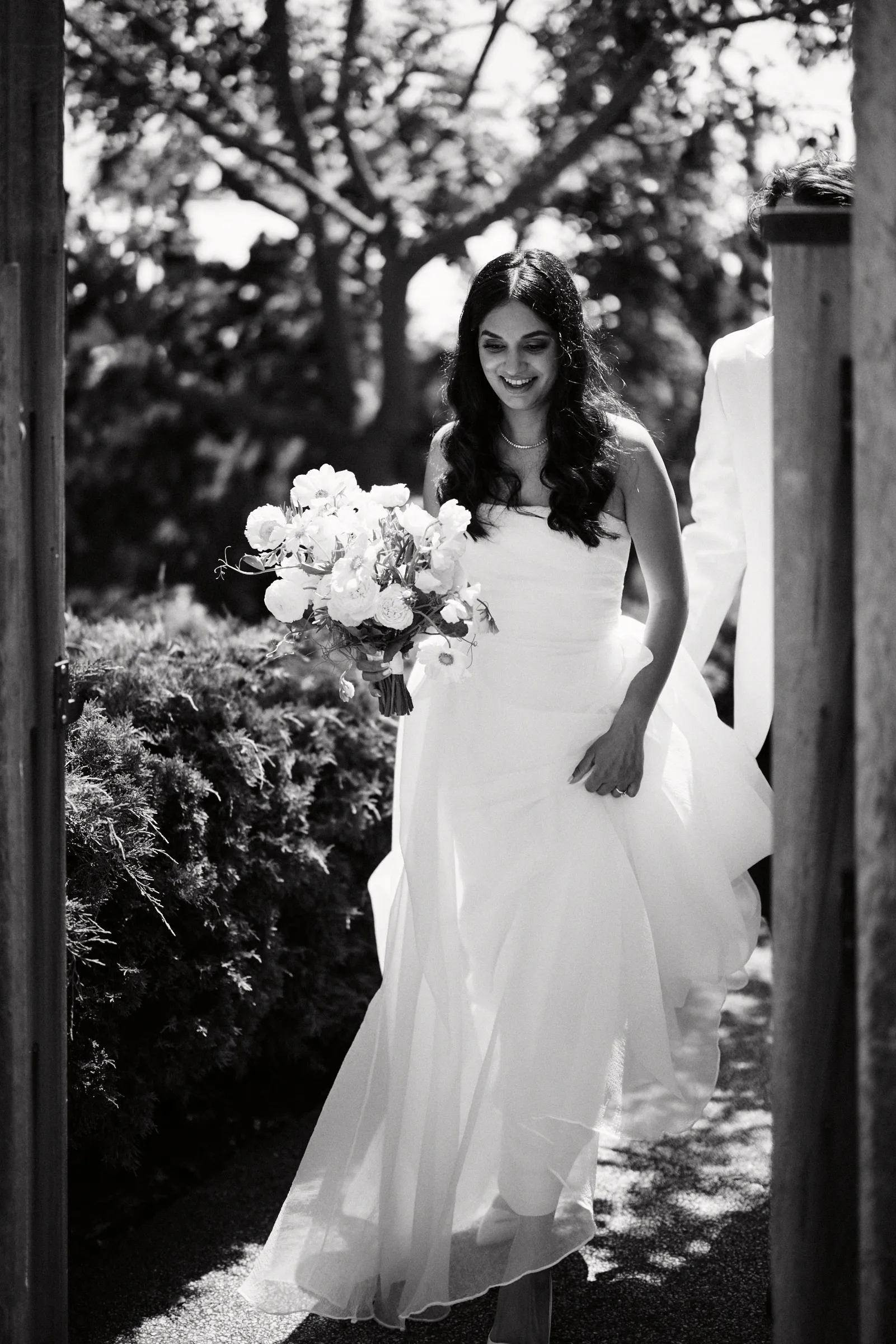 The bride walking under the trees with her bouquet, in black and white