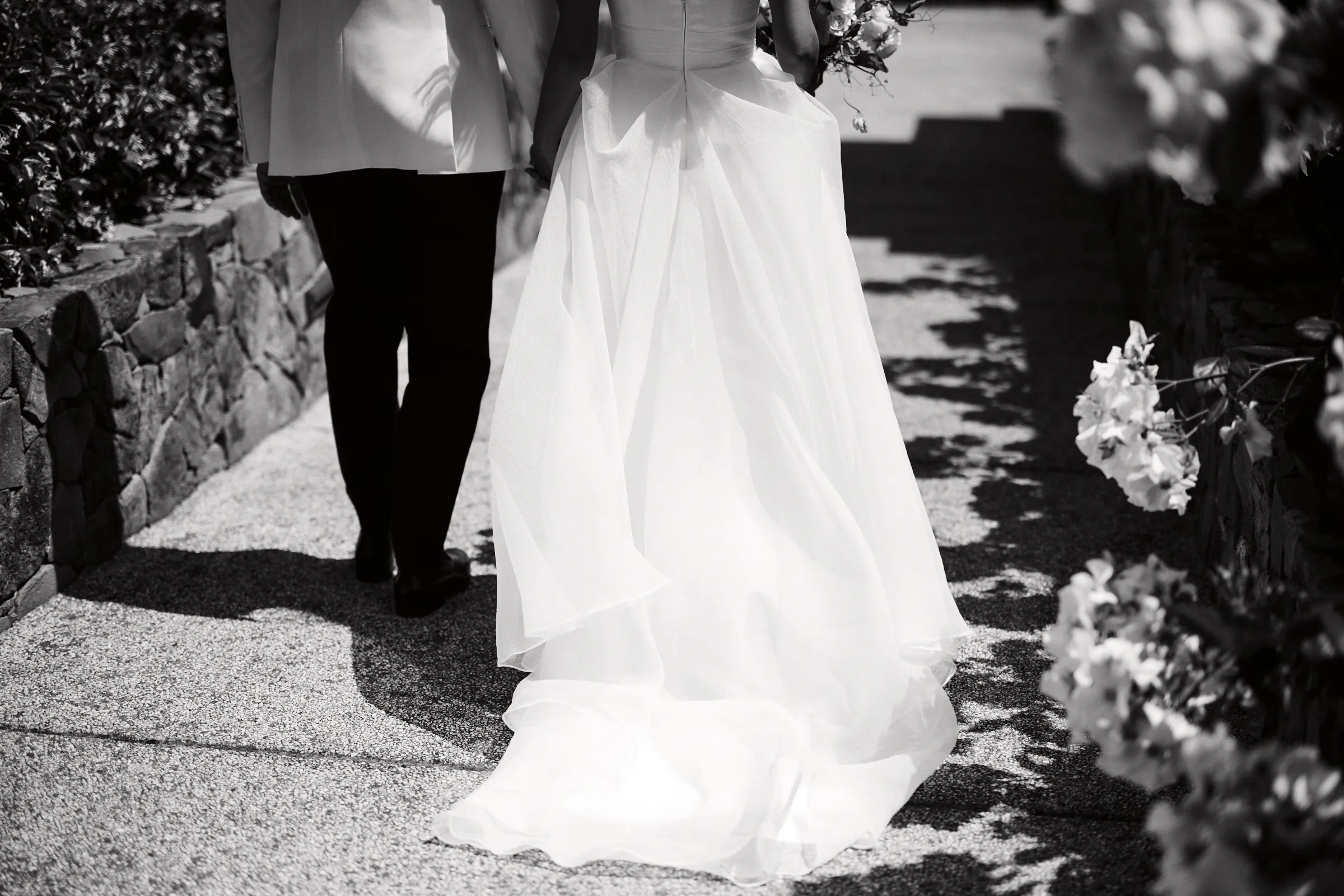 The train of the bride's dress trailing along a stone path, in black and white