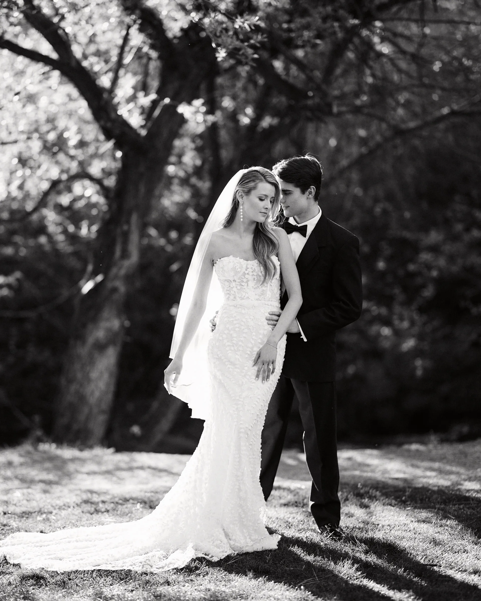 The couple standing close beneath backlit trees, the bride's veil at her shoulder, in black and white