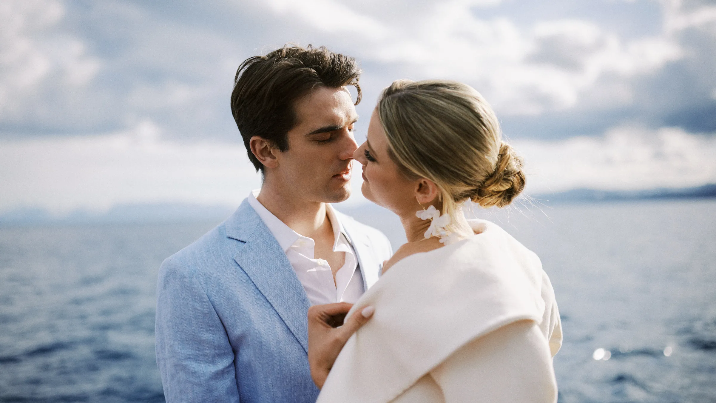 A couple nose to nose on the water, he in a pale blue jacket, she wrapped in ivory