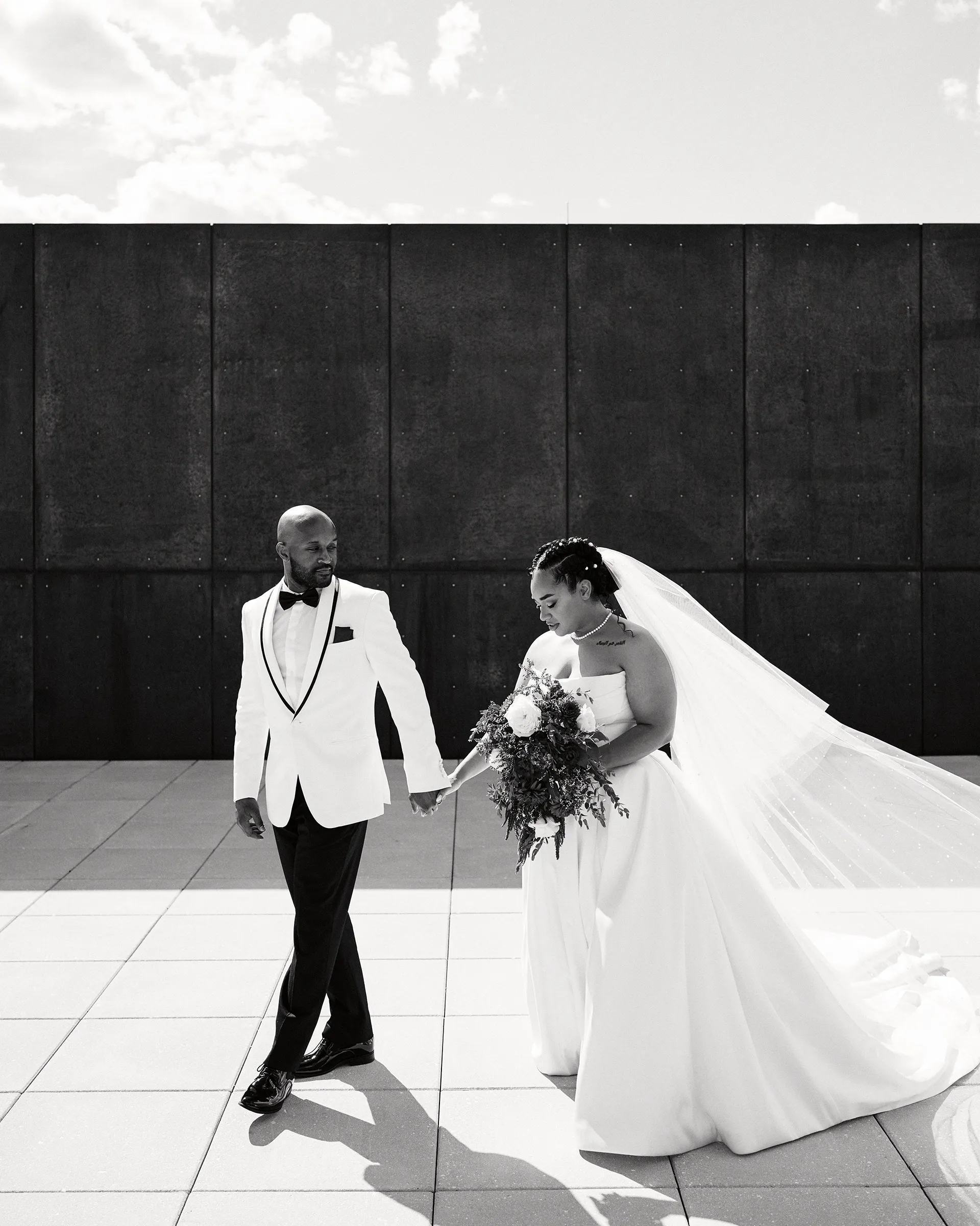 A groom in a white tuxedo jacket leading his bride by the hand across a plaza, in black and white